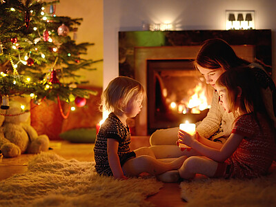 Young mother and her two little daughters sitting by a fireplace holding a candle in a cozy dark living room on Christmas eve
