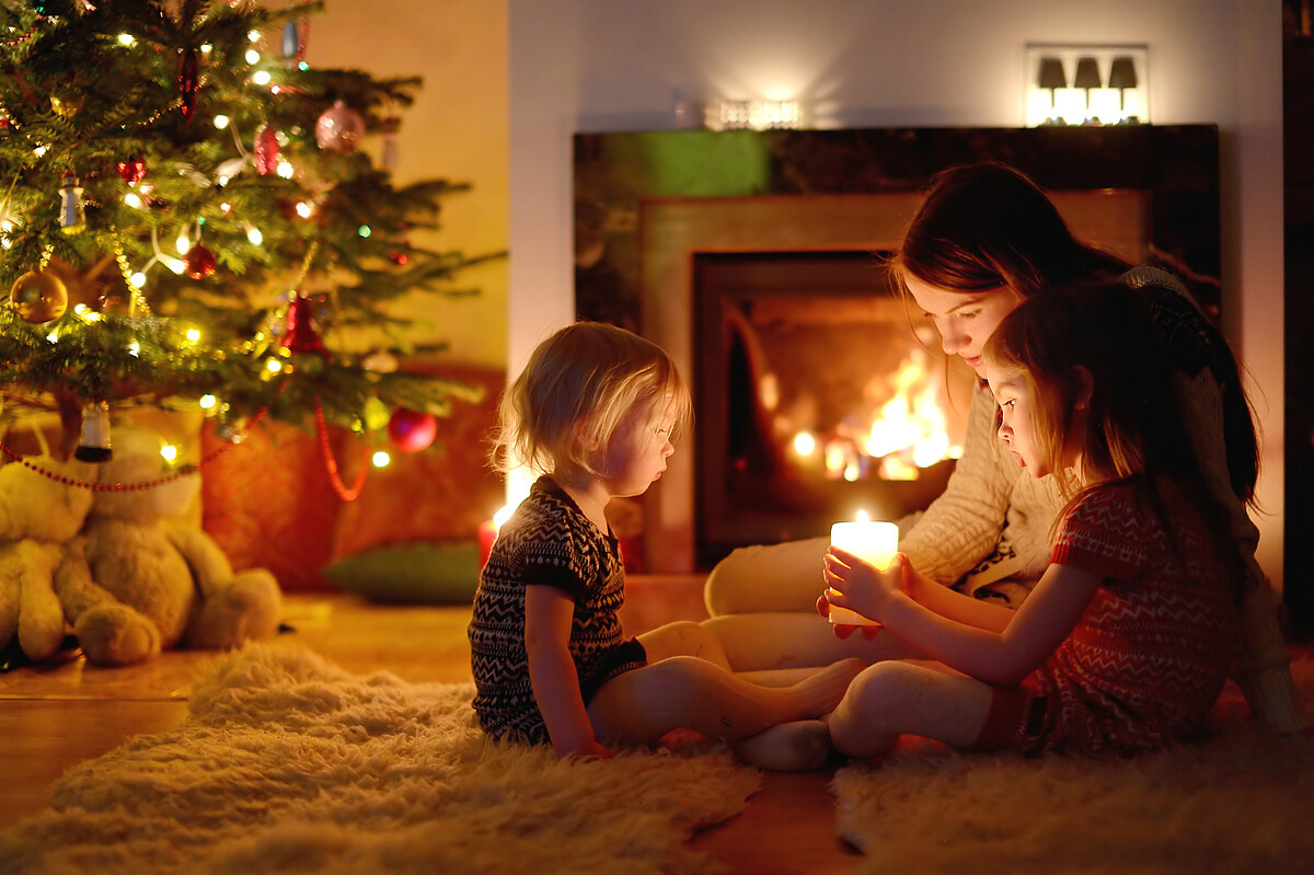 Young mother and her two little daughters sitting by a fireplace holding a candle in a cozy dark living room on Christmas eve