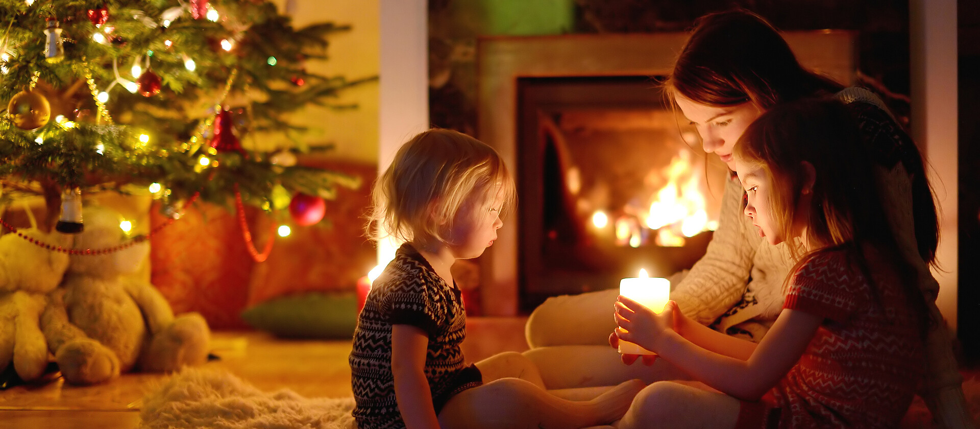 Young mother and her two little daughters sitting by a fireplace holding a candle in a cozy dark living room on Christmas eve